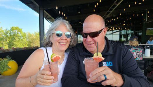 Kent and Becca seated next to each other at a table in a restaurant, each enjoying pink Paloma cocktails!