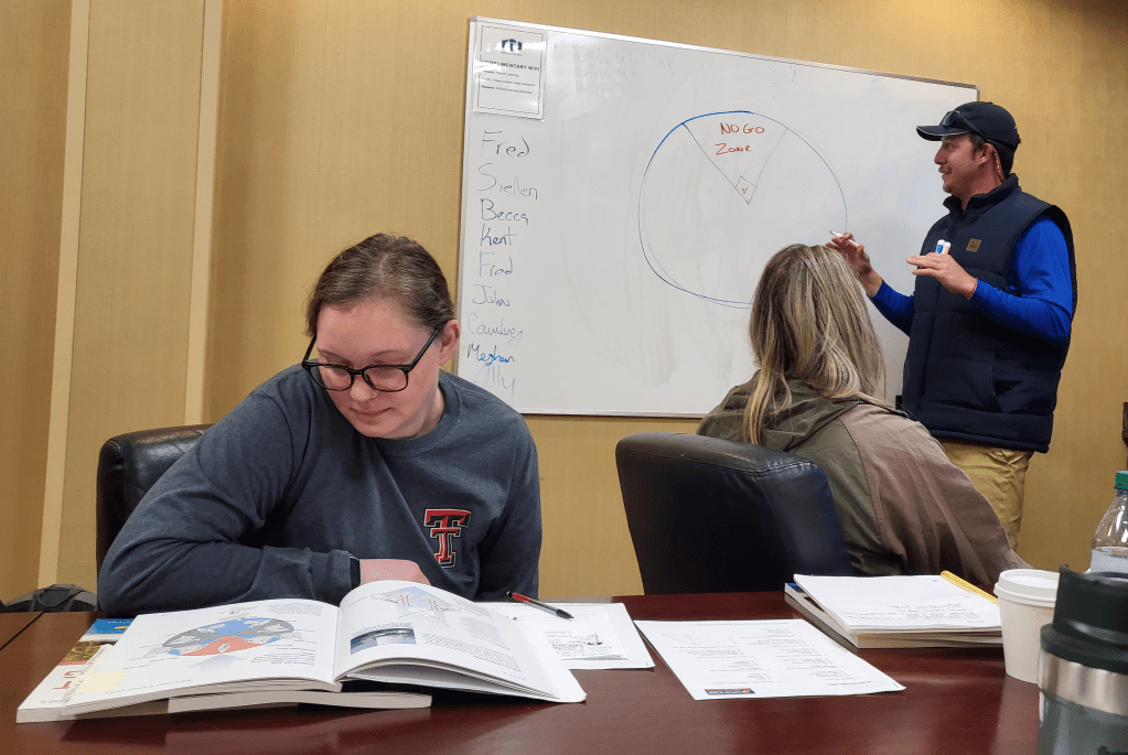 A picture of students sitting at a table while the instructor is standing at a whiteboard, drawing diagrams of the points of sail.