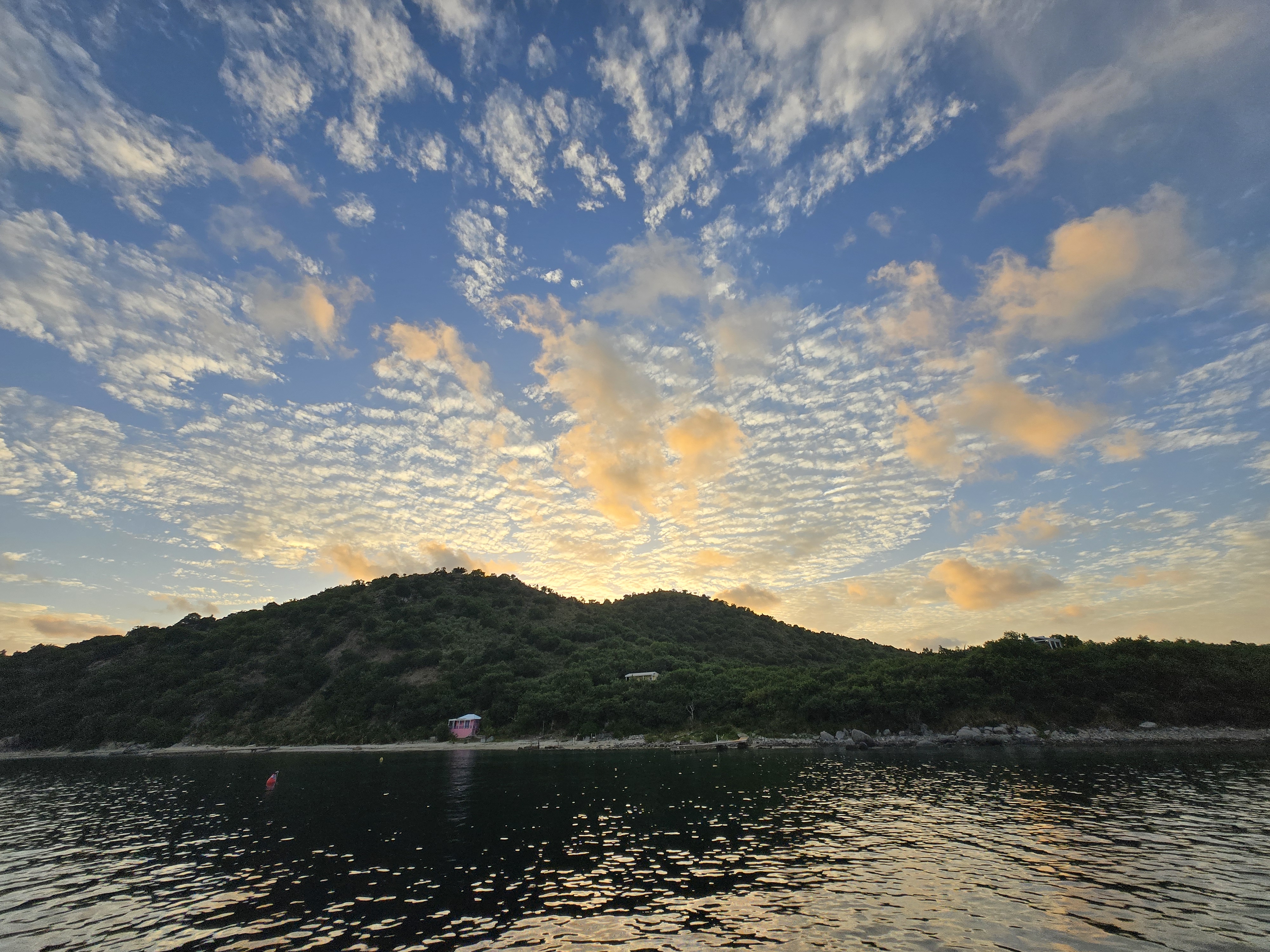 A shoreline, and the sun is coming up behind a hilltop, illuminating the spotty clouds above it