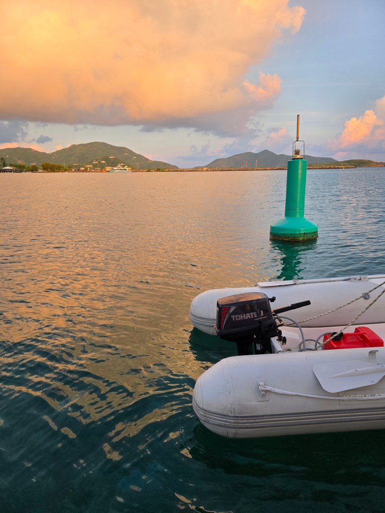 A dinghy floating on the ocean water in the foreground, a bright green bouy in the middle, and islands in the background. 
