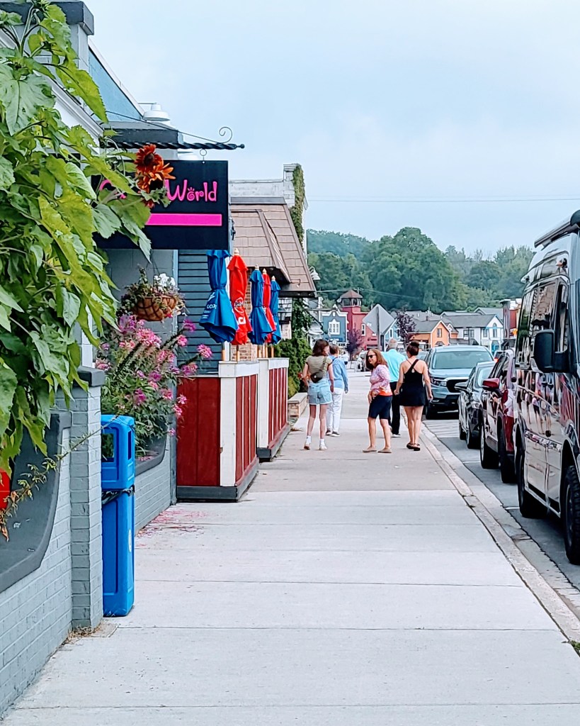 people walking on the sidewalk with storefronts on the left and cars parked on the curb on the right