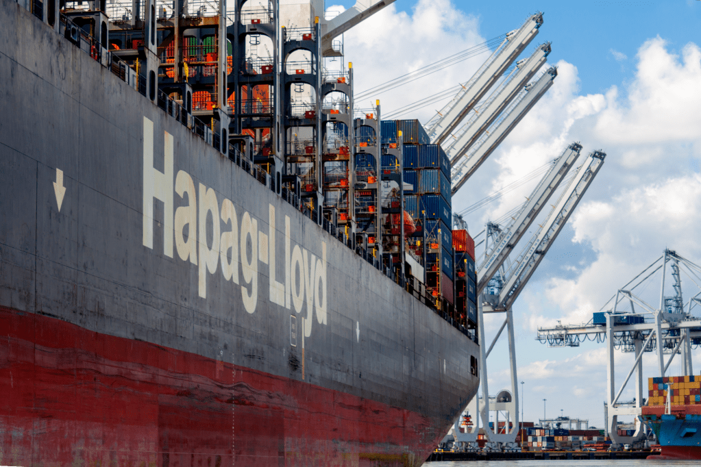 The massive hull of a Hapag-Lloyd container ship, taking on containers, with cranes in the background