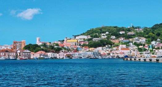 Colorful hillside coastal town with pastel buildings, a yellow‑roofed church tower, and boats docked along the waterfront, set against lush green hills and a clear blue sky.buildings built up on the hillside, with lush trees.