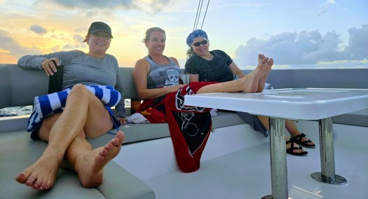 Three people relaxing on a boat at sunset, seated around a white table with cushioned benches, wrapped in towels and blankets, with the ocean and a glowing sky in the background.