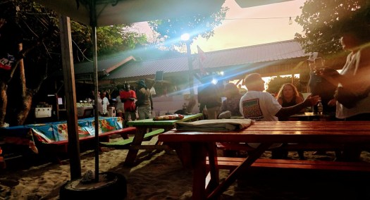 Outdoor beachside dining area at sunset with colorful picnic tables on sand, people gathered around talking and drinking, and a small building with string lights in the background.
