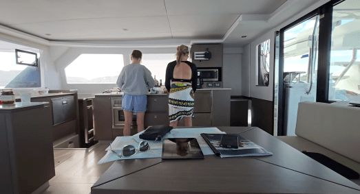 Interior of a modern sailboat galley with two people standing near the kitchen area, talking while preparing food. A dining table in the foreground holds sunglasses, a pouch, a tablet, a magazine, and a nautical chart. Sunlight comes through the windows, illuminating the sleek wood and stainless-steel design.