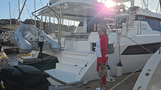 Large white catamaran named “MOORINGS 5000” docked in a marina, with an inflatable dinghy hanging at the stern. A person in a red and white outfit stands on the dock near suitcases and bags under bright sunlight.