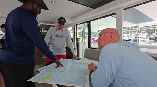 Three people inside a boat reviewing a large nautical chart spread across a table, with one person pointing to a location. Large windows behind them show a marina with docked boats.