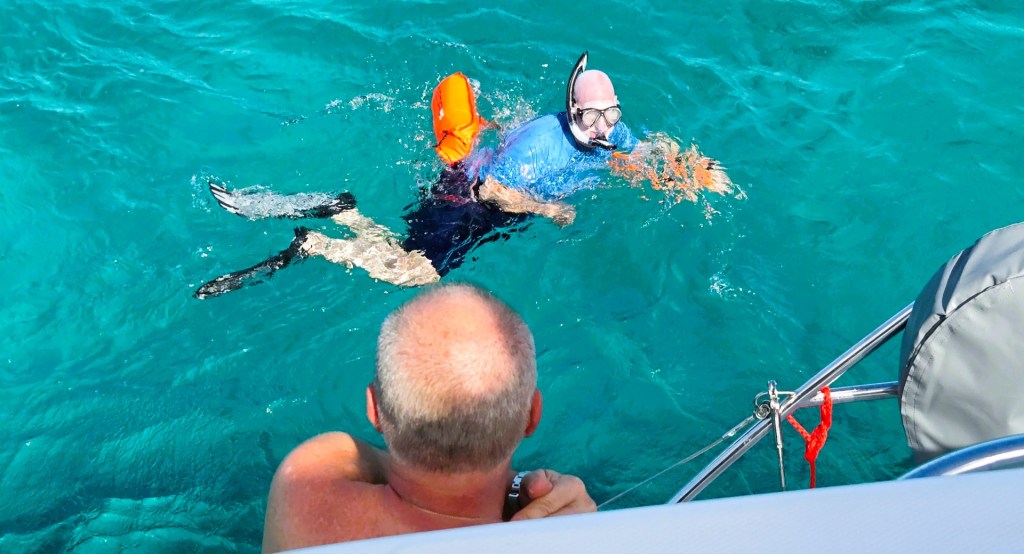 Person snorkeling in clear turquoise water beside a boat, wearing fins, a snorkel mask, and a blue shirt, with an orange flotation device nearby. Another person stands on the boat watching from above.
