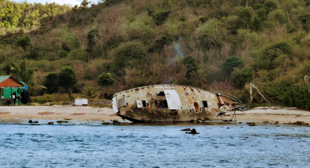 Rusted shipwreck lying on its side on a sandy beach near dense green vegetation, with a small green shack and a person sitting outside beside it.