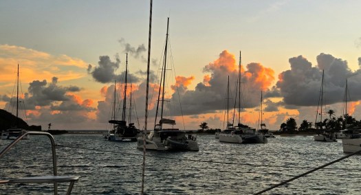 Sailboats and catamarans anchored in calm water at sunset, silhouetted against a sky filled with golden and orange clouds, with palm trees and a small landmass in the background.