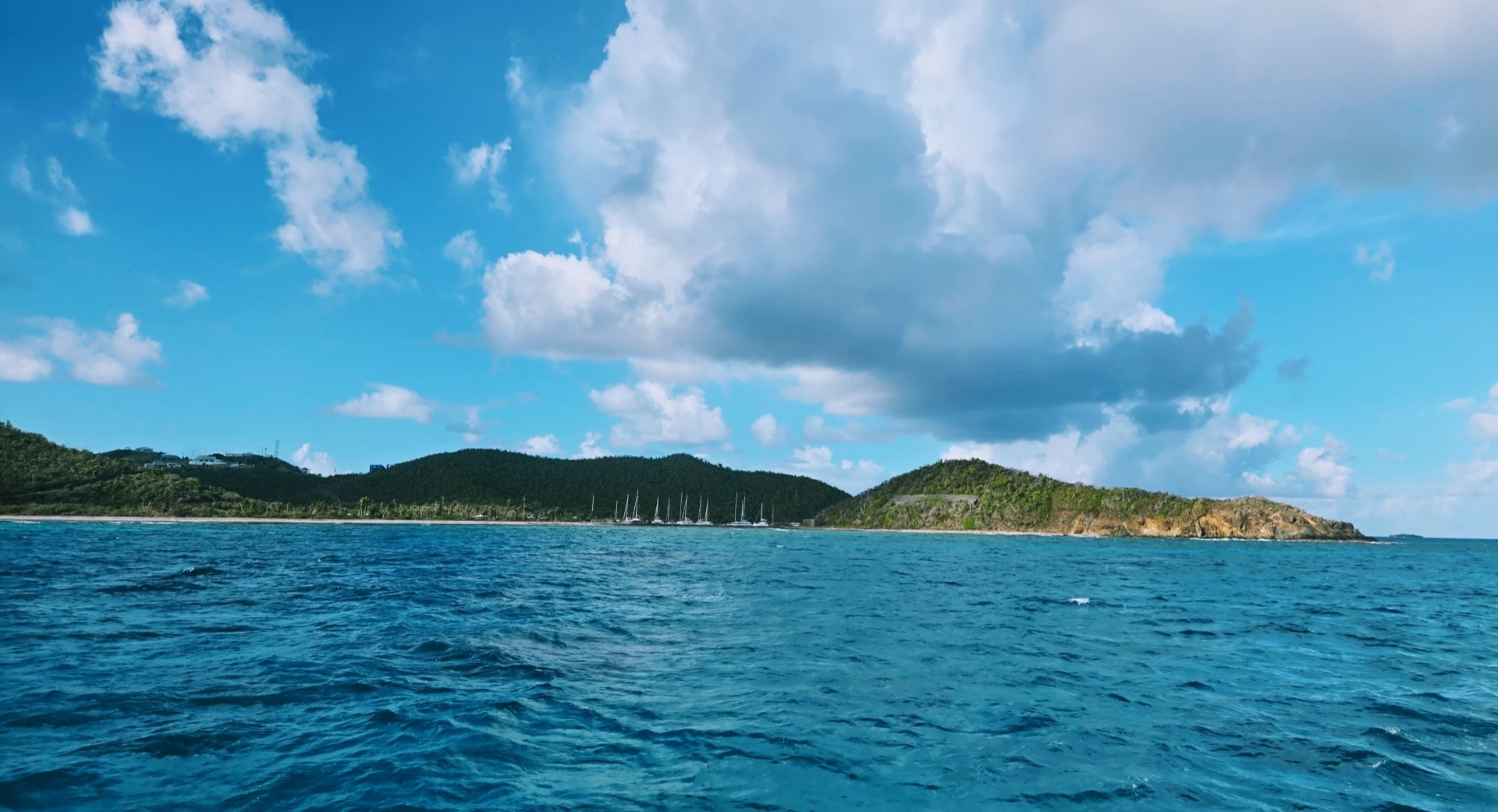 Bright blue ocean in the foreground with a lush green island and a line of sailboats along the shore beneath a mostly clear sky with scattered clouds.