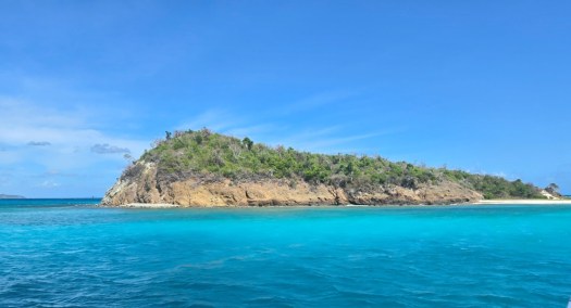 Small lush island surrounded by bright turquoise water, with rocky shorelines and dense green vegetation beneath a clear blue sky.