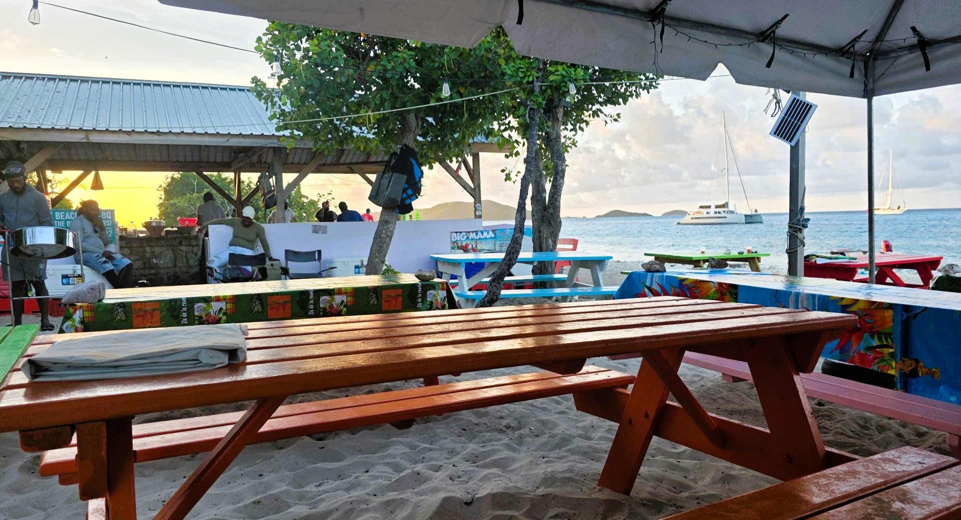 Colorful picnic tables set on a sandy beach under trees and a canopy, with people gathered near a small shelter. Calm ocean water with anchored sailboats and distant islands is visible in the background.