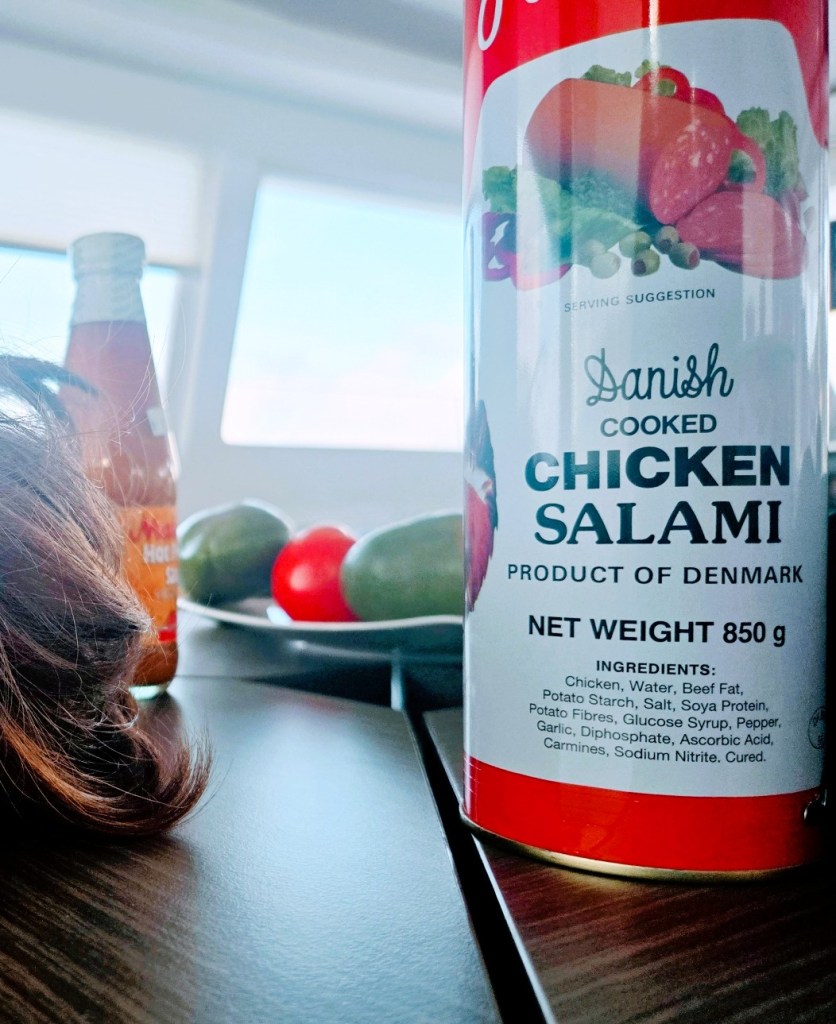 Close-up of a can of Danish cooked chicken salami on a table, with a bottle of hot sauce and a plate of sliced bell peppers in the background. Part of a person’s hair is visible on the left side.