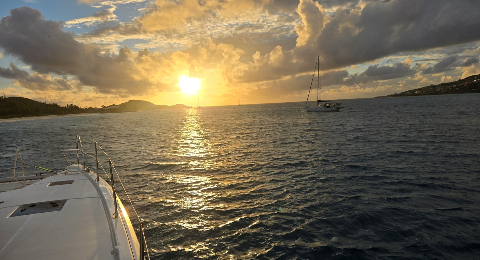 Boat bow in the foreground facing a golden sunset over calm ocean water, with a sailboat anchored mid‑distance and hilly coastline on both sides beneath dramatic, glowing clouds.