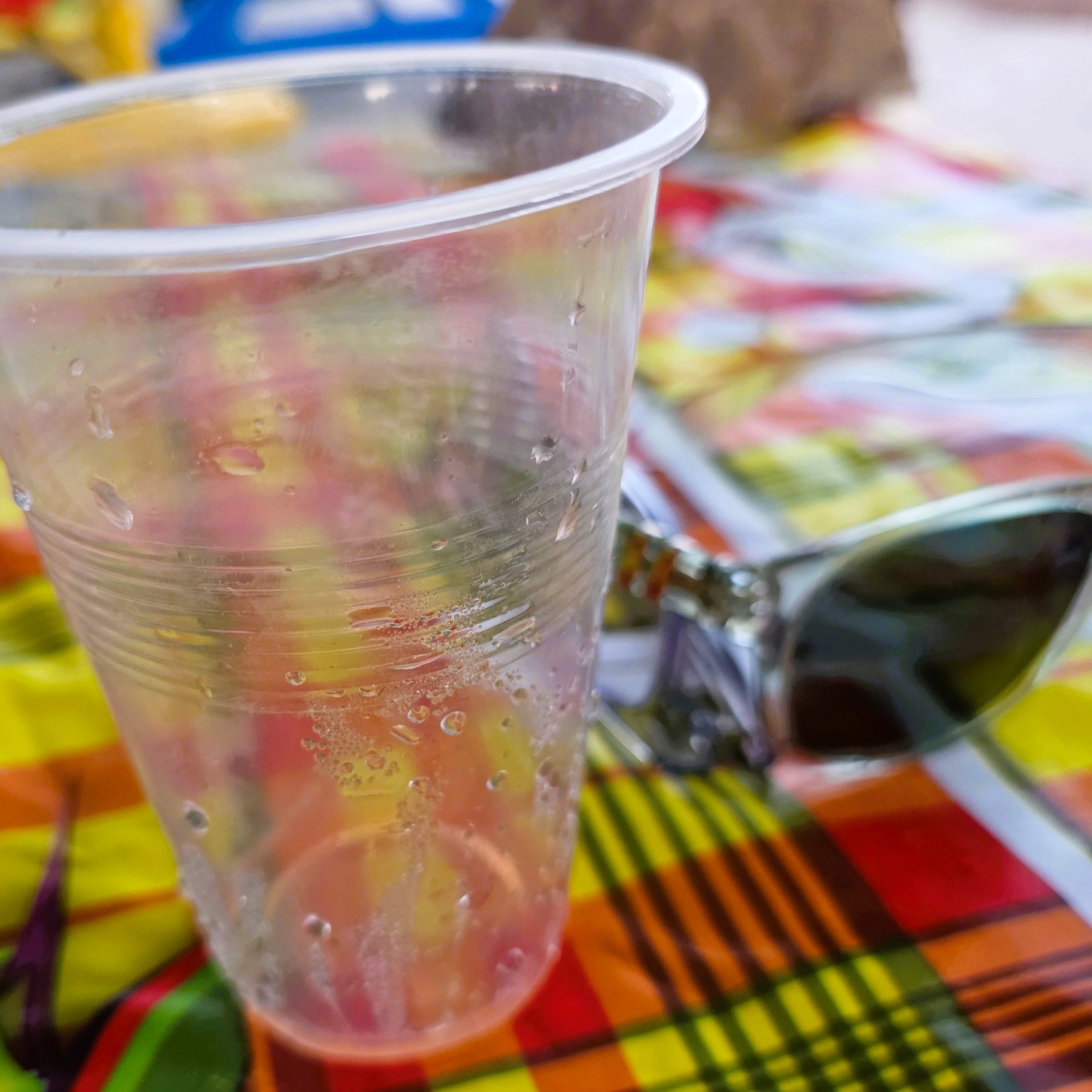 Close-up of a clear plastic cup with condensation on a colorful checkered tablecloth, with sunglasses resting behind it and blurred picnic items in the background.
