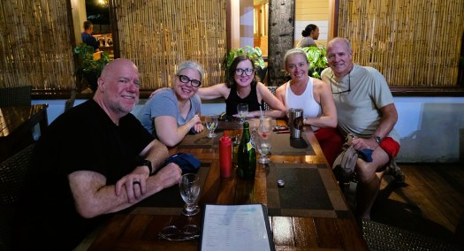 Five people sitting around a wooden table in a casual restaurant with bamboo screens and plants in the background, smiling as they enjoy drinks and conversation. Glasses, a bottle, a red condiment bottle, a stainless-steel tumbler, a menu, and eyeglasses are on the table.