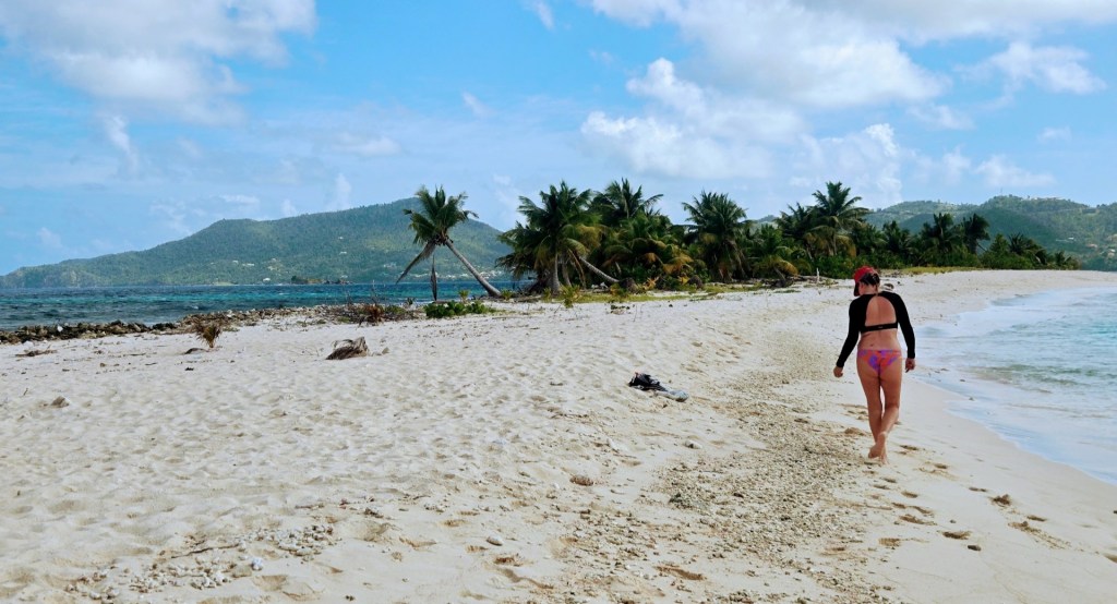 Person walking along a white-sand tropical beach beside clear blue water, with palm trees lining the shore and green hills rising in the background. They wear a red cap, a black long-sleeve top, and pink-and-purple swimwear.