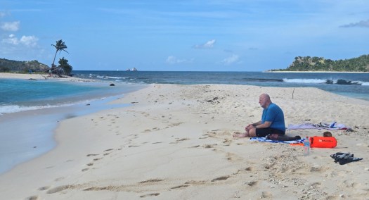 Man sitting on a sandy tropical beach with snorkeling gear, an orange dry bag, and a water bottle nearby. Clear turquoise water, small islands, and leaning palm trees appear in the background under a mostly sunny sky.