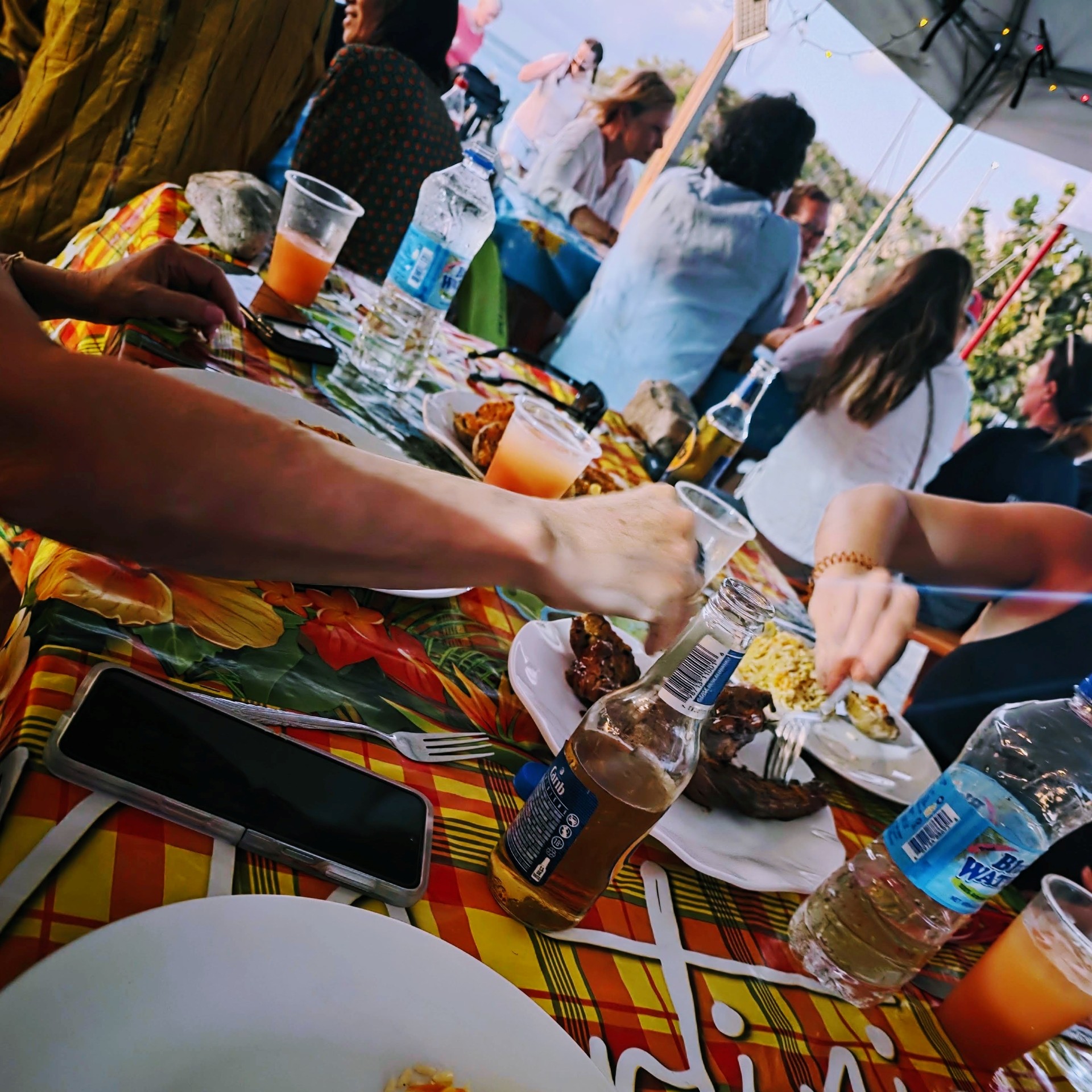 People gathered around a table covered with a colorful floral and plaid tablecloth, eating plates of food and drinking bottled water, beer, and orange beverages, with more people talking under a canopy lit by string lights in the background.