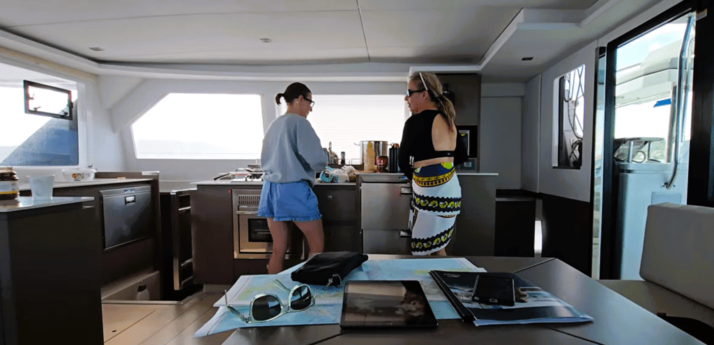 Interior of a modern sailboat galley with two people standing near the kitchen area, talking while preparing food. A dining table in the foreground holds sunglasses, a pouch, a tablet, a magazine, and a nautical chart. Sunlight comes through the windows, illuminating the sleek wood and stainless-steel design.