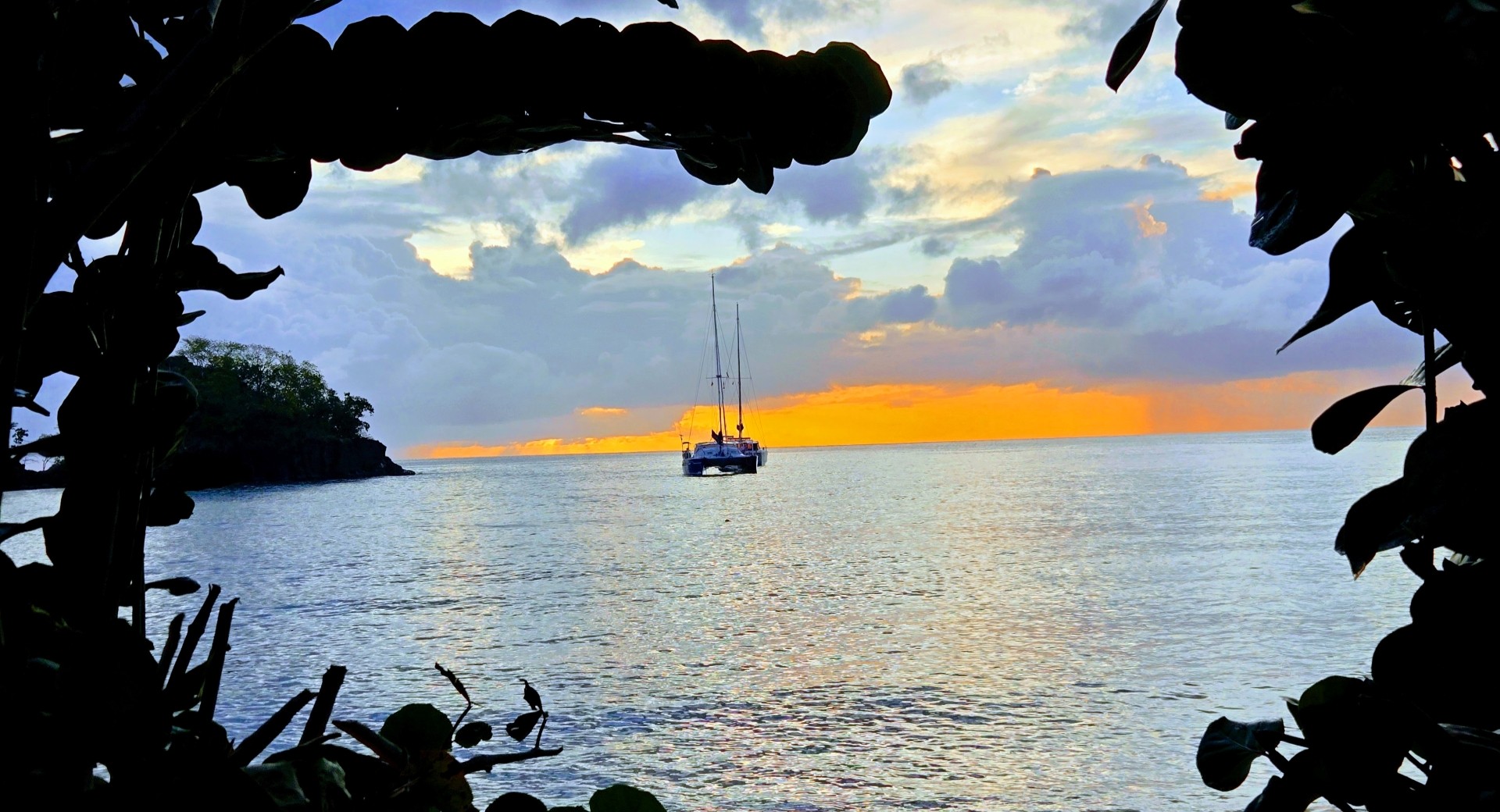 Sunset view of a sailboat anchored on calm water, seen through a tunnel of green foliage framing a sandy beach.