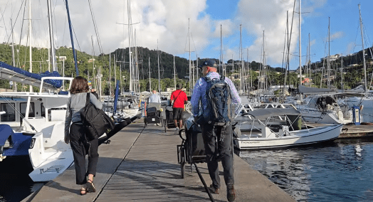 Four people walking down a wooden pier in a marina lined with sailboats and yachts, two of them pulling carts loaded with luggage. Green hills with scattered houses rise in the background under a partly cloudy sky.