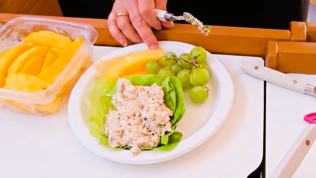 An overhead close-up shot of a plate that contains tuna salad cupped in a large lettuce leaf, green grapes, and slices of yellow mango. 