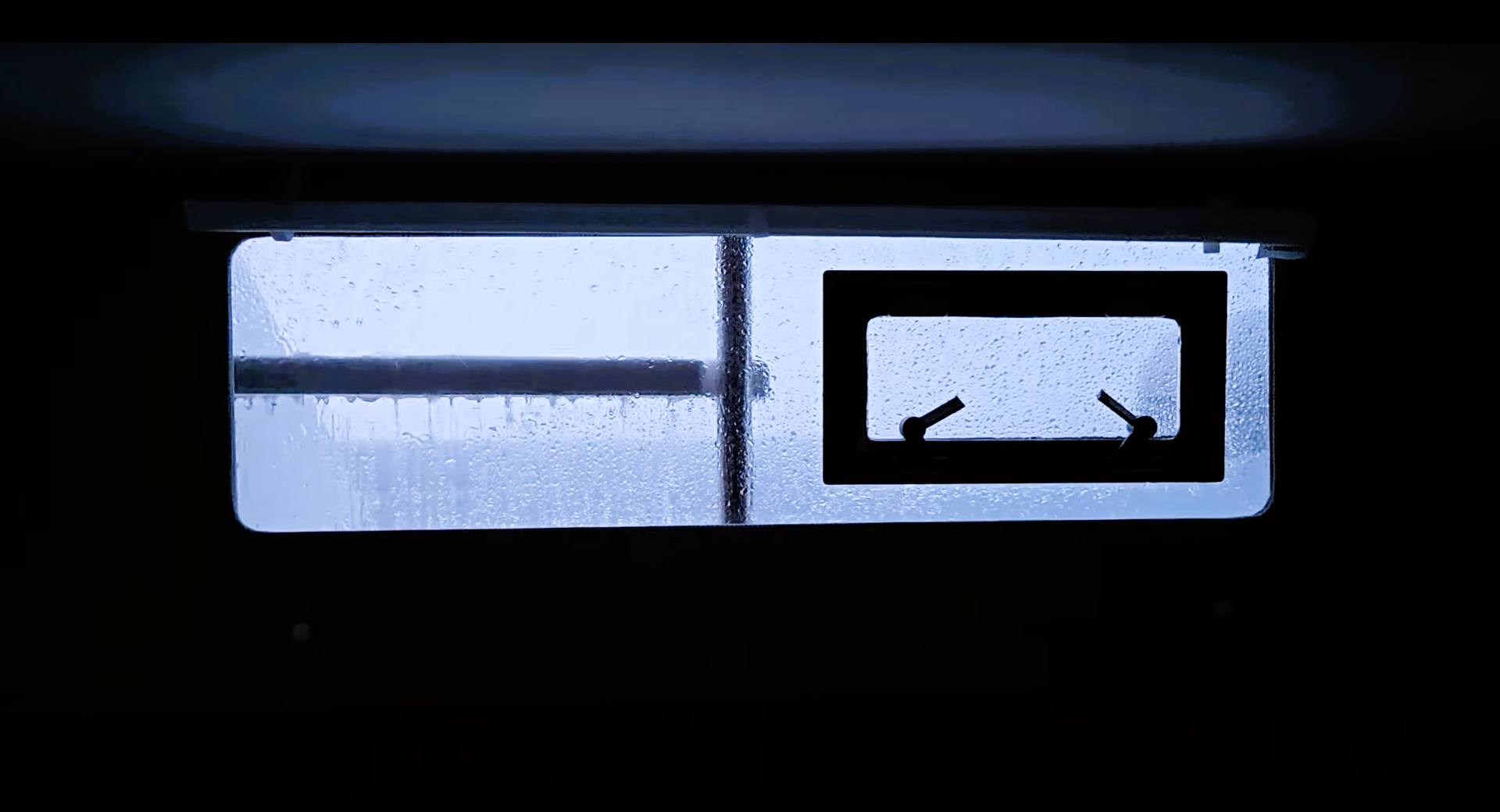 Rain‑covered window with a hatch secured by two locking handles, seen from indoors with soft blue light reflecting on the ceiling.