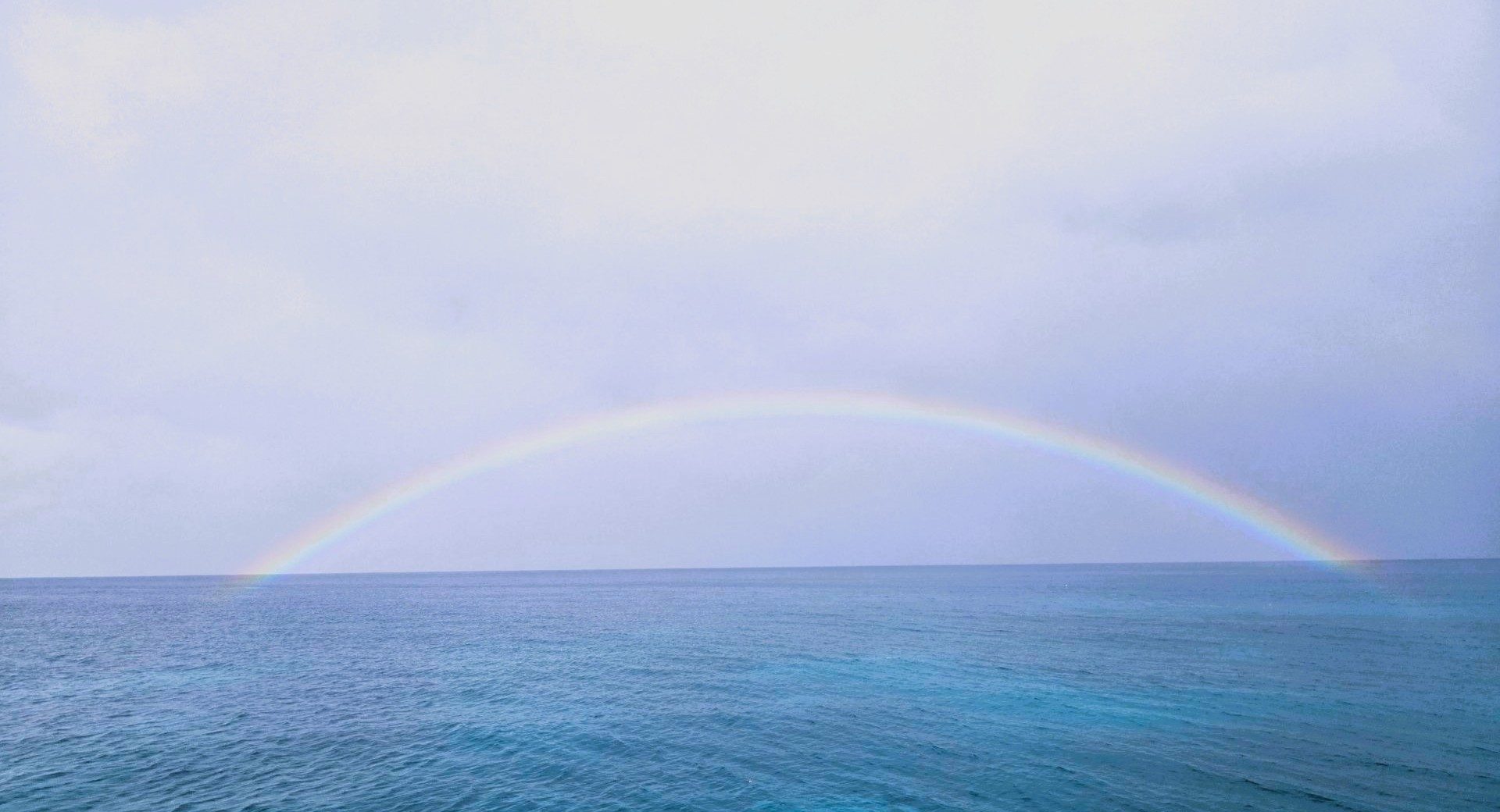 Bright rainbow arching across an overcast sky above a calm ocean with gentle waves.